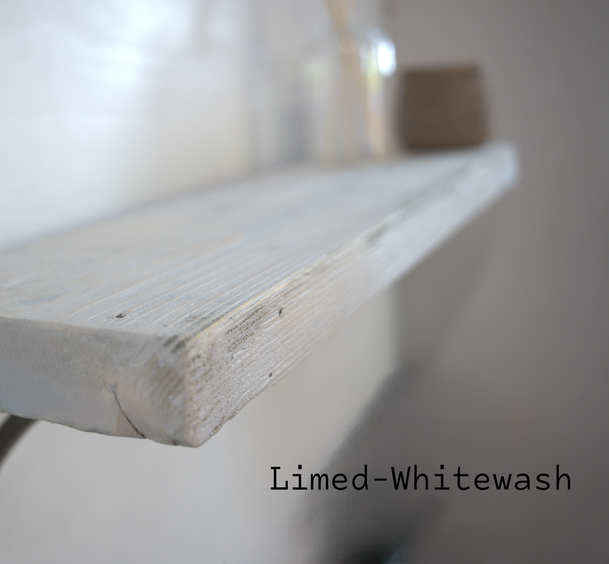 A close-up of a whitewashed wooden shelf with a distressed, lime-washed finish, showing the wood grain and chipped edges. The shelf is mounted against a neutral wall with decorative items blurred in the background.