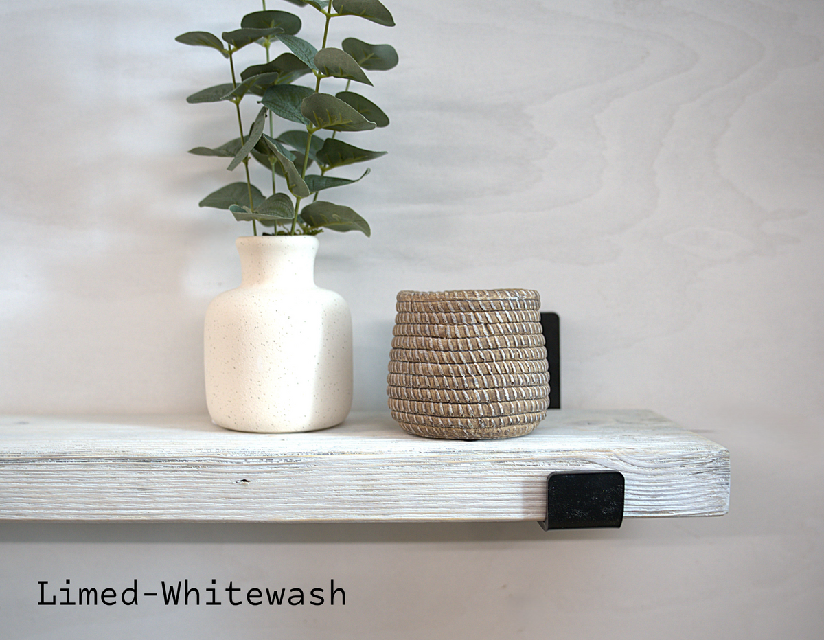 A rustic white wooden floating shelf mounted on a light gray wall, styled with a textured off-white ceramic vase holding green eucalyptus stems and a small woven natural-fiber basket. Black metal brackets secure the shelf to the wall.
