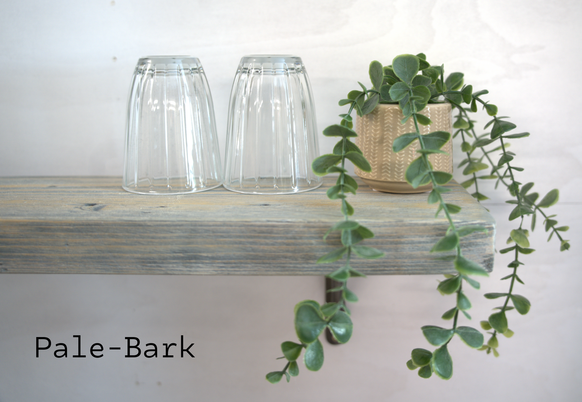Two clear glass cups upside down on a rustic pale-bark wooden shelf, with a trailing green plant in a woven gold pot to the right. The text 'Pale-Bark' is visible in the lower-left corner.