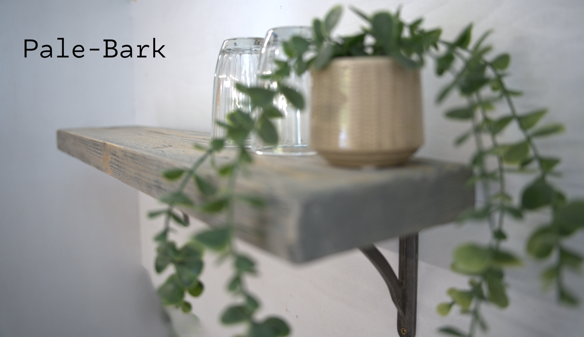 A rustic floating shelf made of pale-bark wood with a dark metal bracket, styled with a small potted trailing green plant and two clear drinking glasses against a white wall.