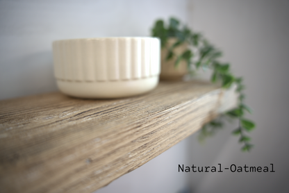 A small cream-colored ceramic bowl with a fluted exterior sits on a rustic wooden shelf, with a blurred green trailing plant in the background. The text 'Natural-Oatmeal' is printed in the bottom right corner.