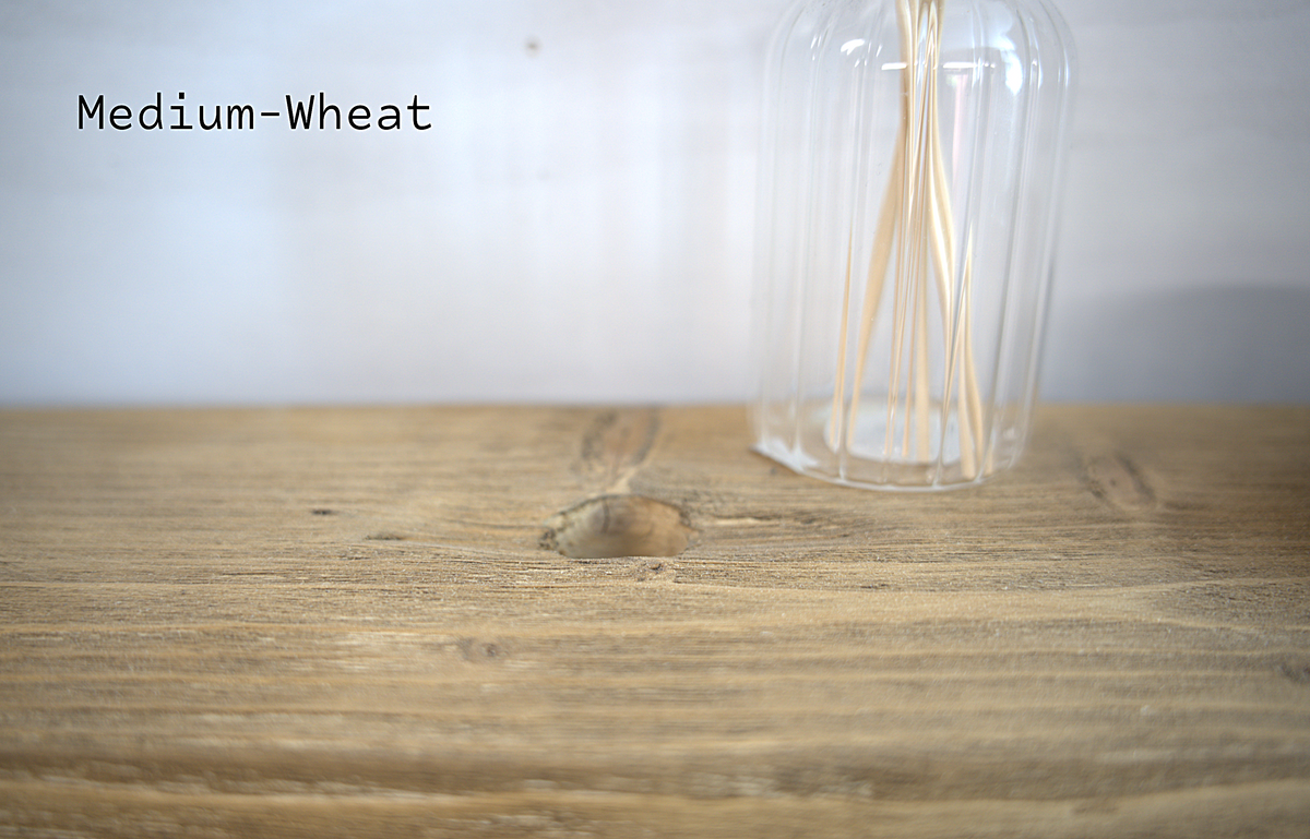 A close-up of a medium-wheat colored wood surface with a clear ribbed glass bottle containing thin wooden sticks, against a light neutral background. The text 'Medium-Wheat' is visible in the upper left.