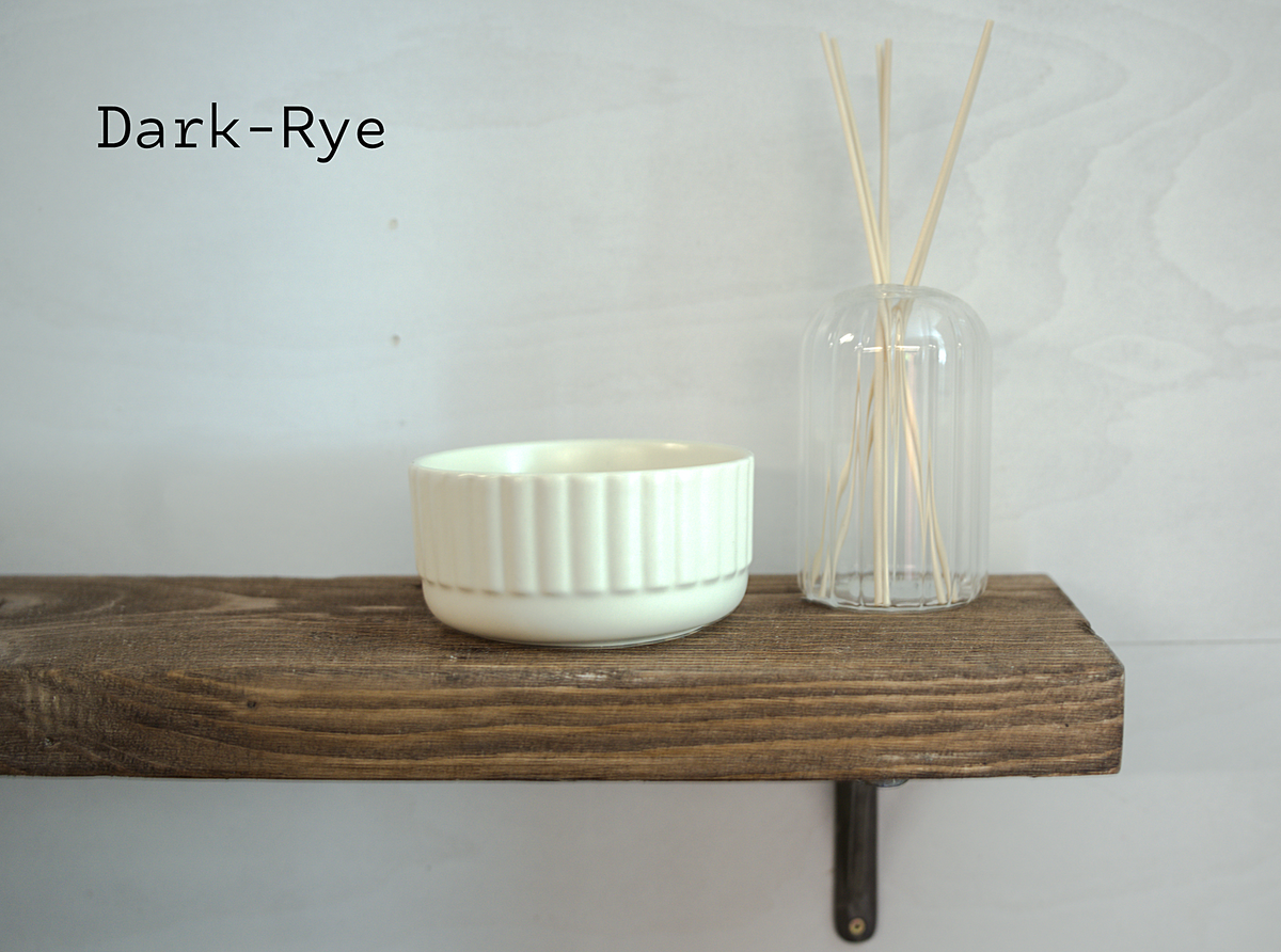 A dark-rye scented candle in a ribbed white ceramic container displayed on a rustic wooden shelf next to a clear glass reed diffuser with wooden sticks. The text 'Dark-Rye' is printed in black in the upper left corner.